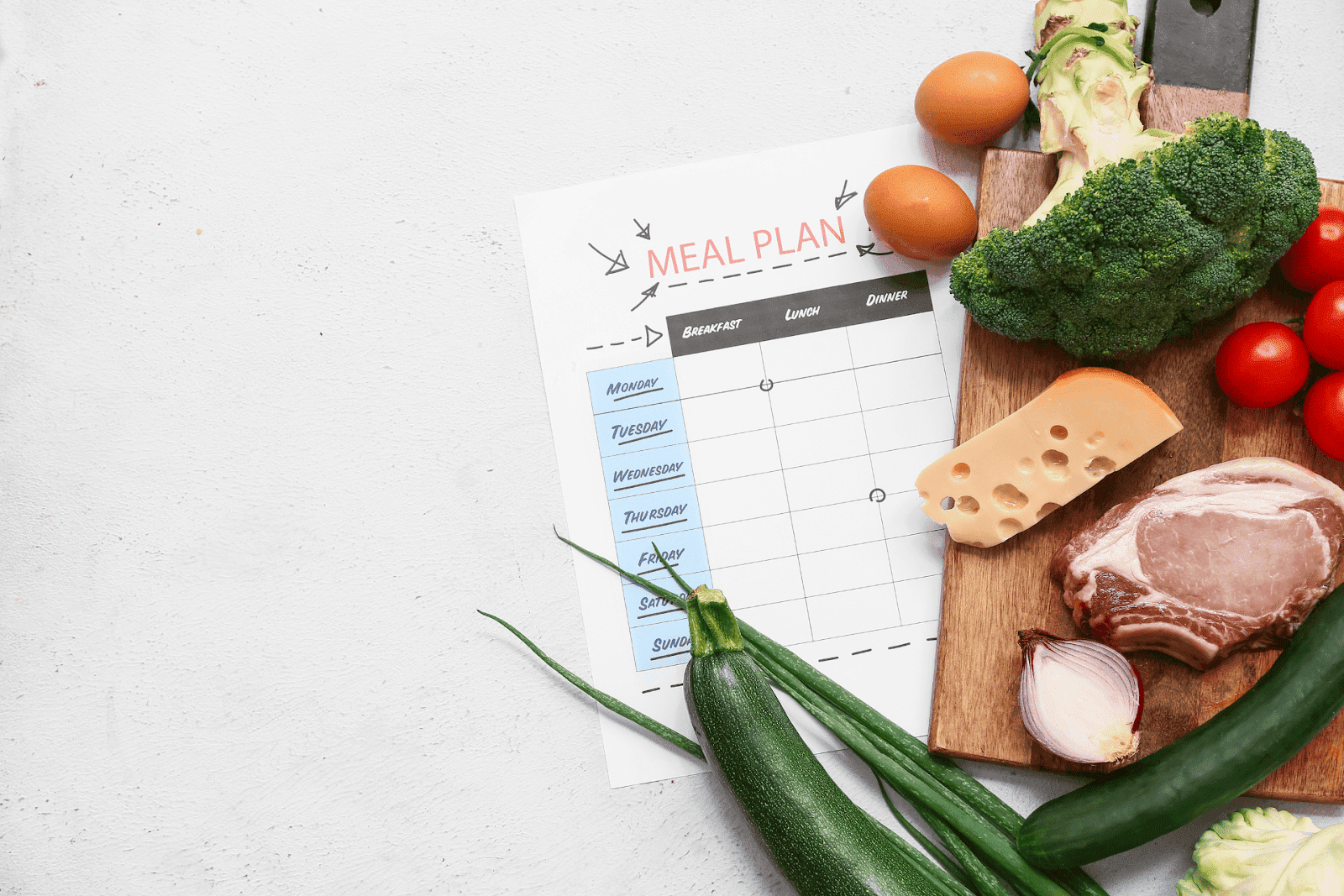 Weekly meal plan sheet next to fresh vegetables, eggs, cheese, and meat on a cutting board