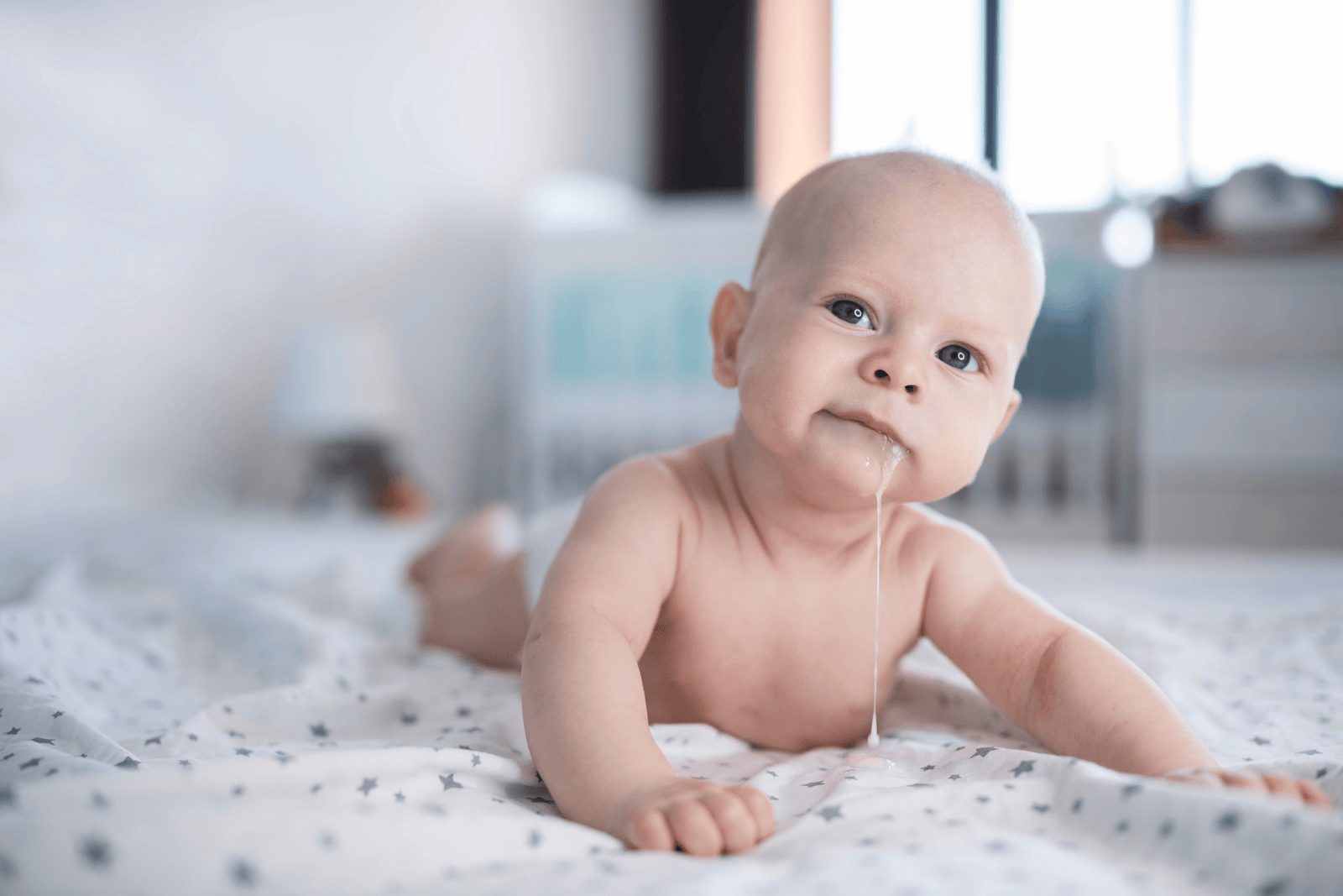 A baby lying on their stomach on a soft bed, looking toward the camera with a small amount of milk dribbling from their mouth, illustrating infant reflux or spit-up in a calm home setting.