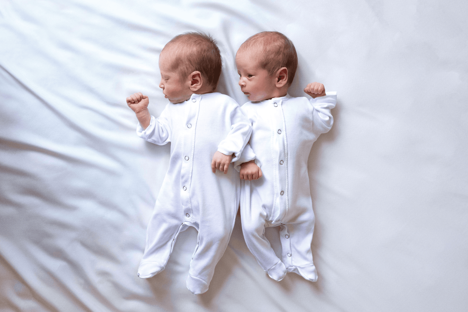 Newborn twin babies lying side by side in white outfits.