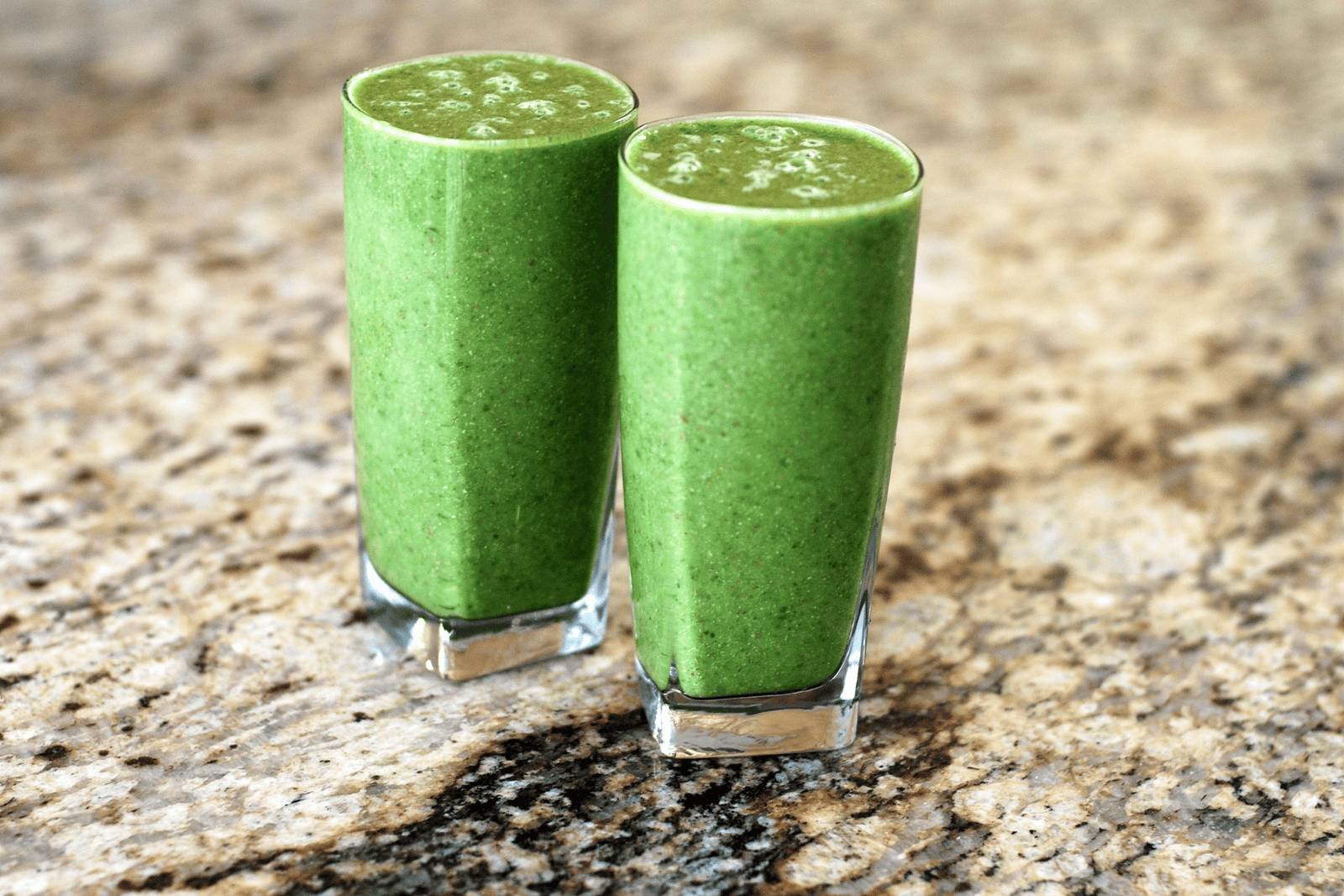 Two tall glasses of green smoothies placed on a kitchen countertop.