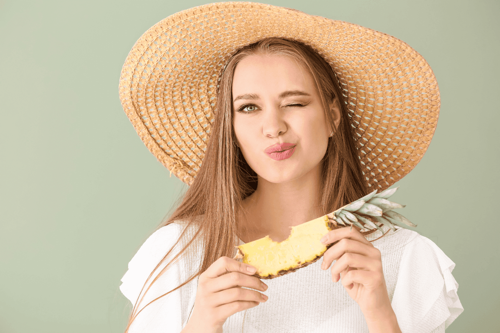 Woman wearing a straw hat winking while holding a slice of pineapple.