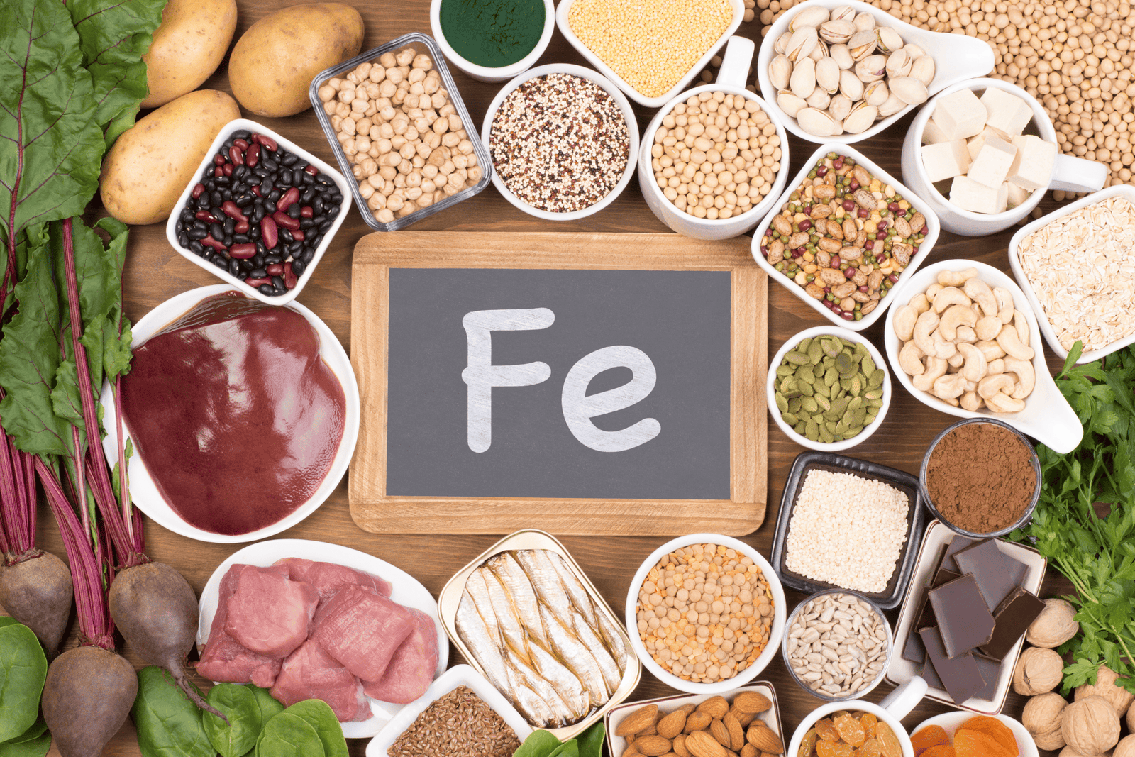 Iron-rich foods arranged around a chalkboard reading \"Fe,\" including leafy greens, beans, nuts, seeds, tofu, fish, and meat.