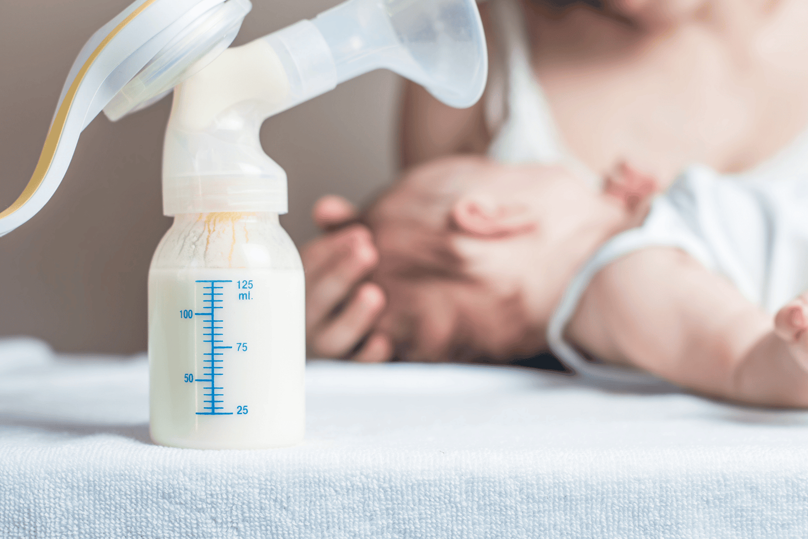 Breast pump bottle filled with milk in the foreground, with a mother holding her newborn baby softly blurred in the background.