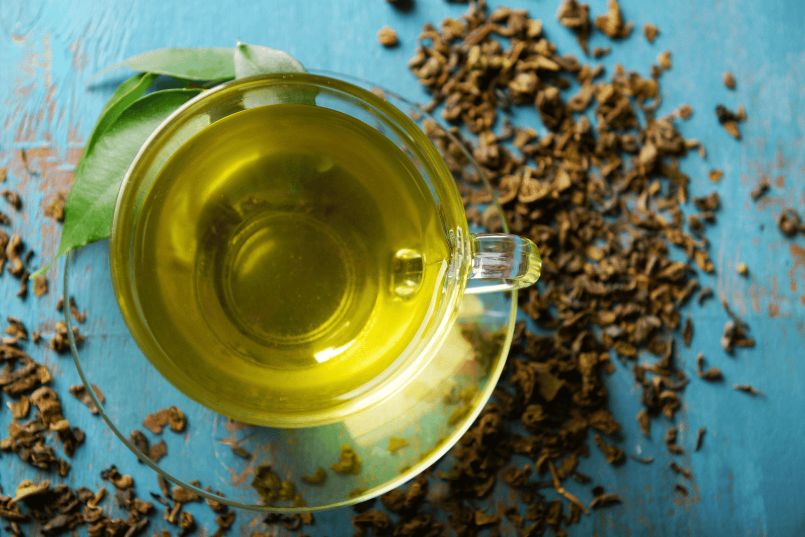 Top-down view of a cup of green herbal tea in a glass mug on a blue surface, surrounded by loose dried tea leaves and a few fresh green leaves.