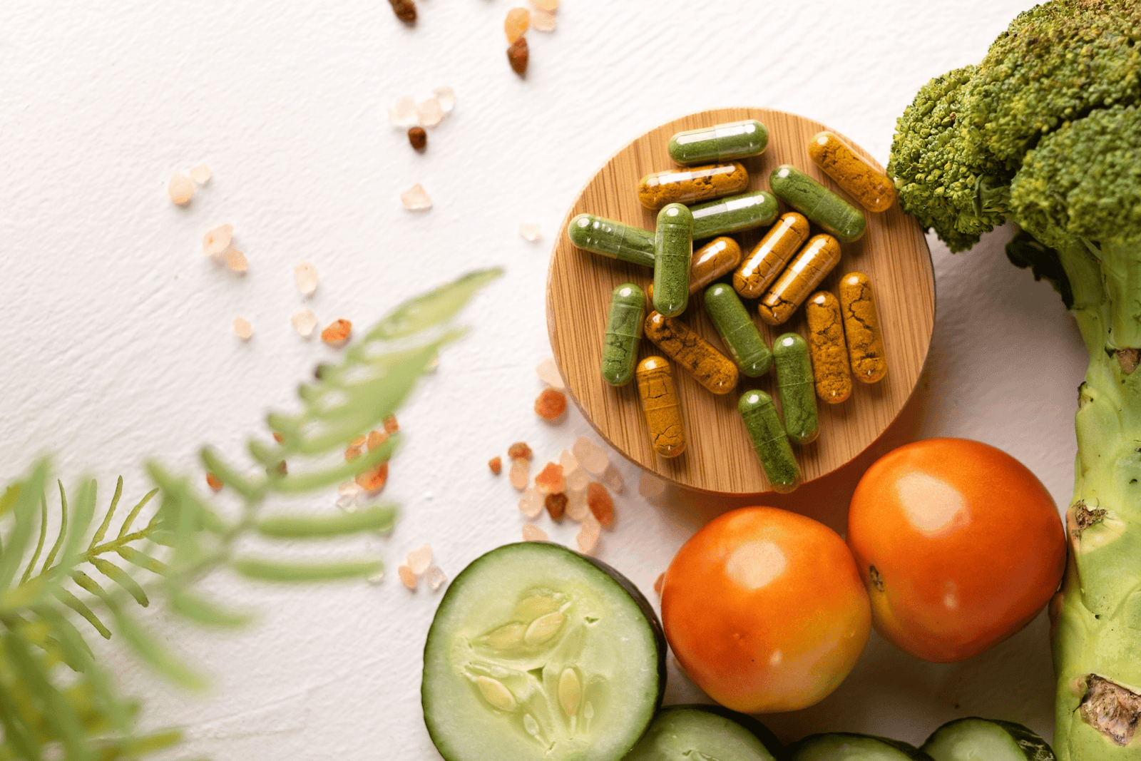 Green and yellow supplement capsules on a wooden dish surrounded by fresh vegetables on a white background.